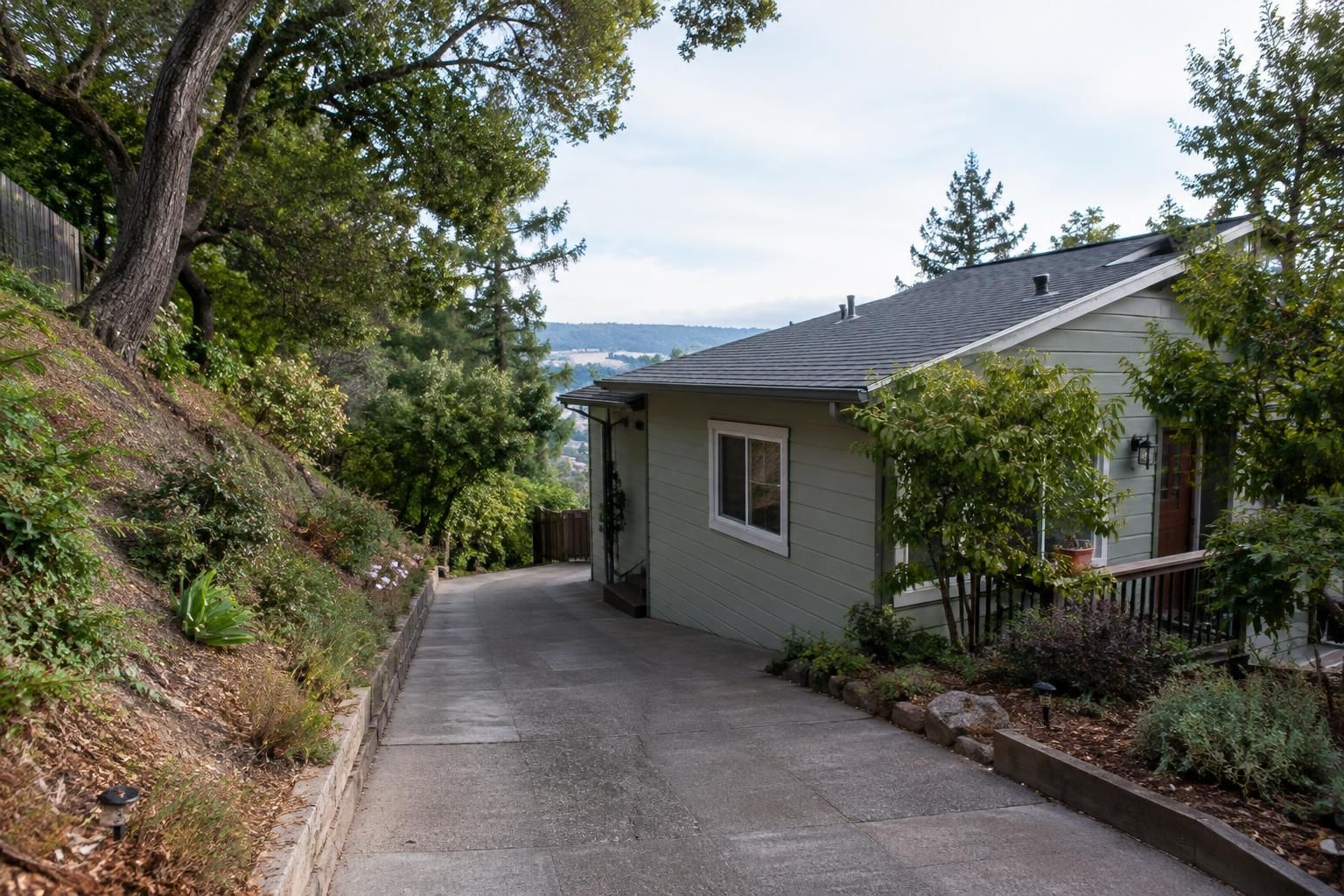 Narrow driveway beside a hillside home with surrounding trees and sloped landscaping.